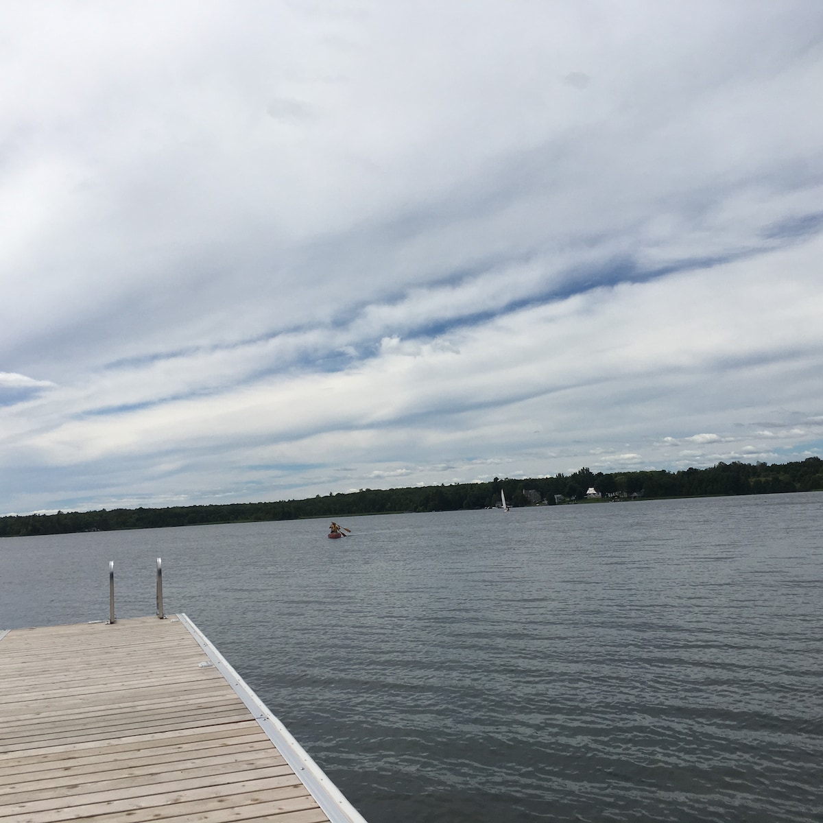 A wooden dock extends over calm waters, with a view of a peaceful lake. Light clouds drift across the sky, reflecting a sense of tranquility. In the distance, a kayak can be seen gliding over the water, with tree-lined shores framing the scene.
