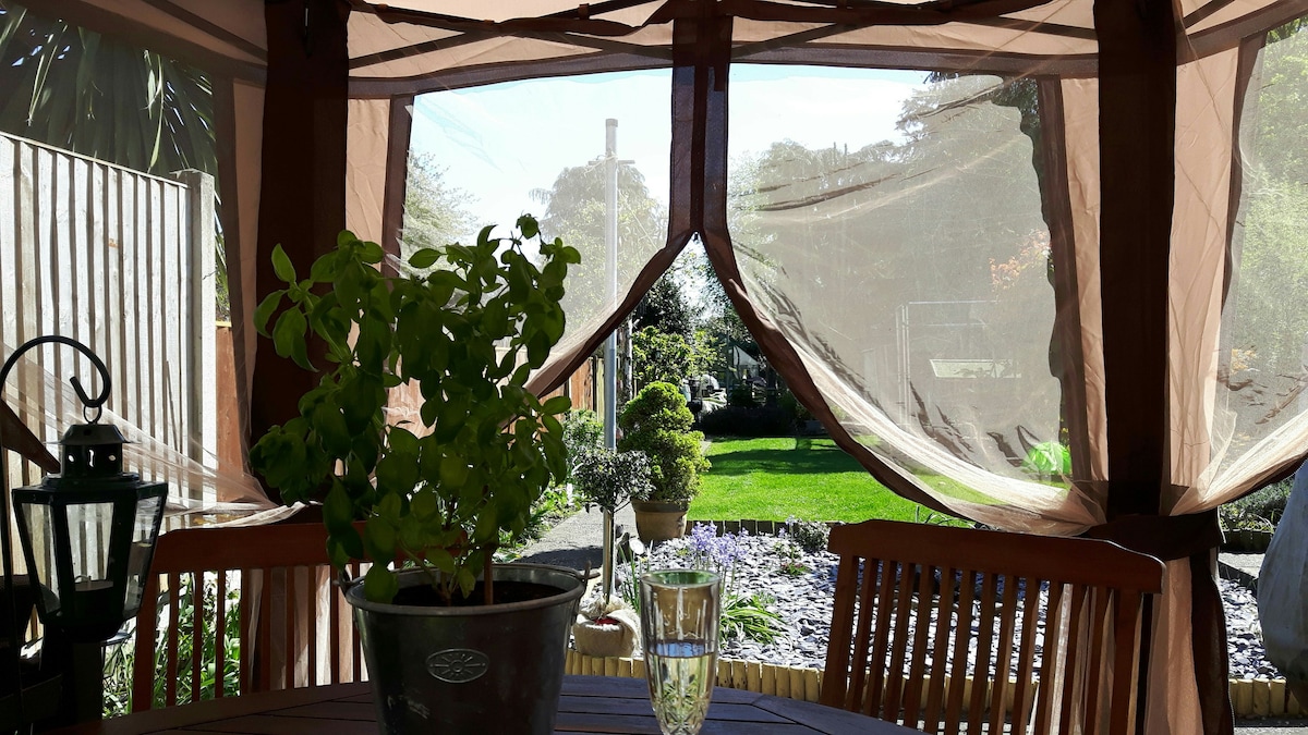 A serene garden setting is visible through a tent structure, framed by soft, flowing fabric. Garden features include neatly trimmed shrubs and a vibrant lawn, with a small table and chairs in the foreground. Natural light filters through, enhancing the peaceful ambiance.