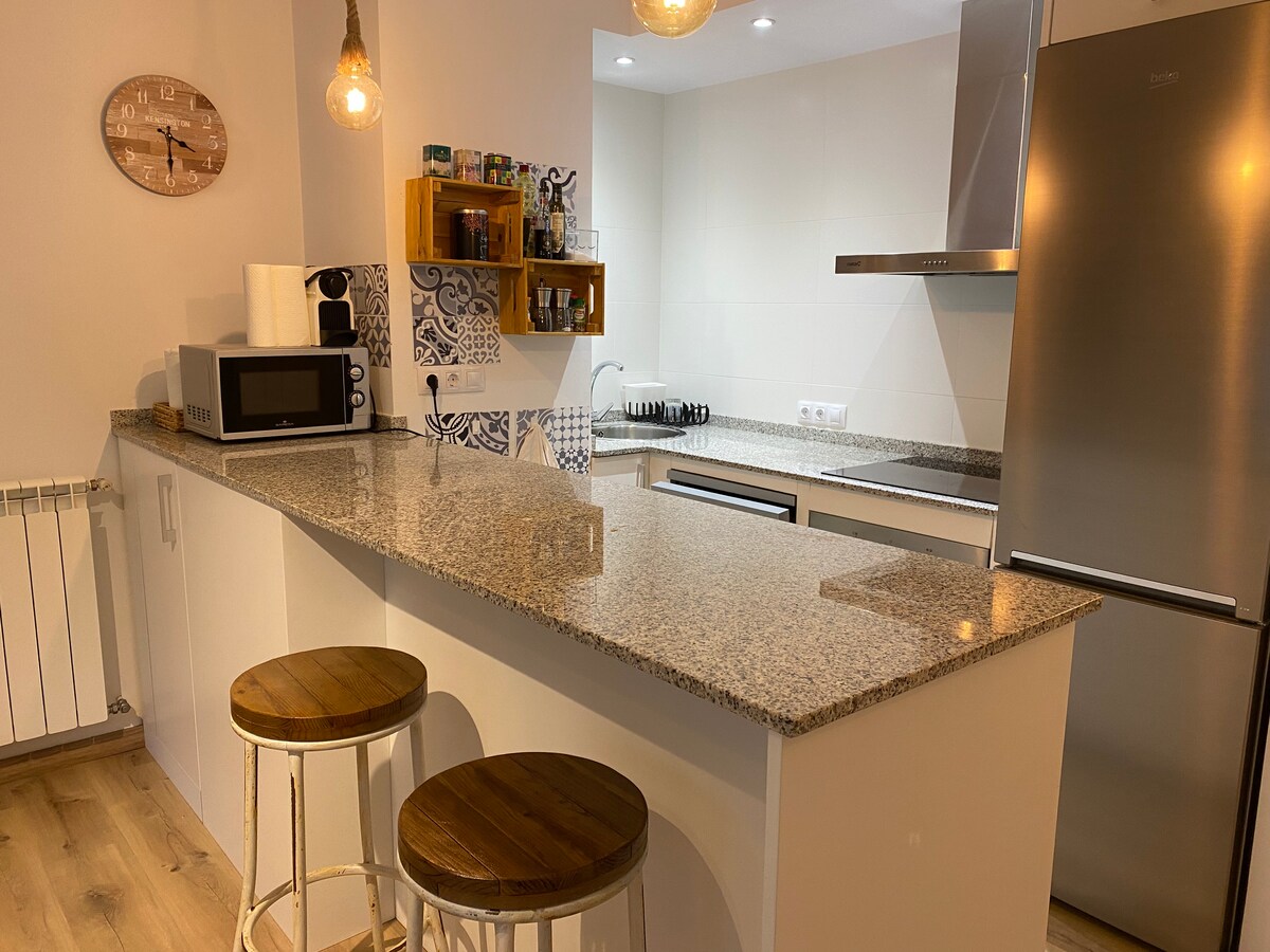 A well-equipped kitchen is visible, featuring modern appliances including a microwave and refrigerator. The granite countertop is complemented by two wooden stools. Open shelving displays various kitchen items, and a wall clock adds a functional touch. Soft lighting enhances the welcoming ambiance.