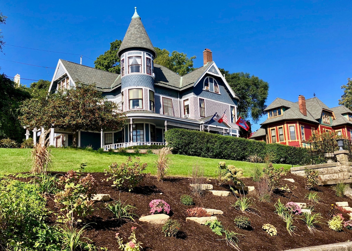 A historic Queen Ann Victorian house is set against a blue sky, featuring a turret and varied architectural details. A well-maintained lawn with landscaped flower beds enhances the exterior, while flags gently flutter from the porch. Surrounding trees add natural beauty to the scene.
