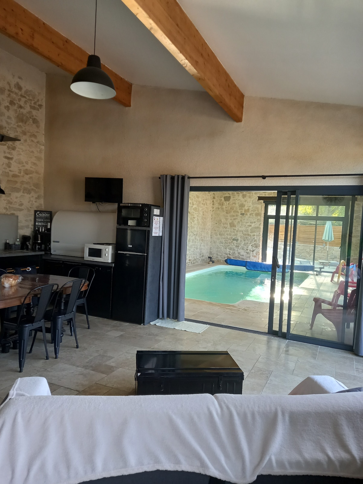 A spacious living area features high wooden beams and a light-toned stone wall. It opens through large glass doors to a covered pool area. A dining table with black chairs and a kitchenette equipped with essential appliances, including a microwave and refrigerator, are visible.