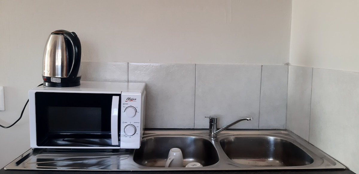 A kitchen area features a stainless steel sink with a single faucet and a microwave situated below. A silver kettle rests on the counter, providing additional utility. Light-colored tiles cover the wall behind the appliances, contributing to a clean and functional space.