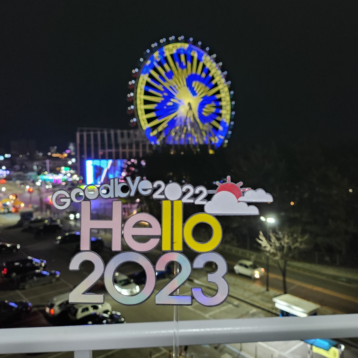 A vibrant Ferris wheel illuminated in blue and yellow lights stands in the background, while a decorative sign reading 'Goodbye 2022 Hello 2023' is positioned in the foreground. The scene captures the lively atmosphere of the surroundings at night.