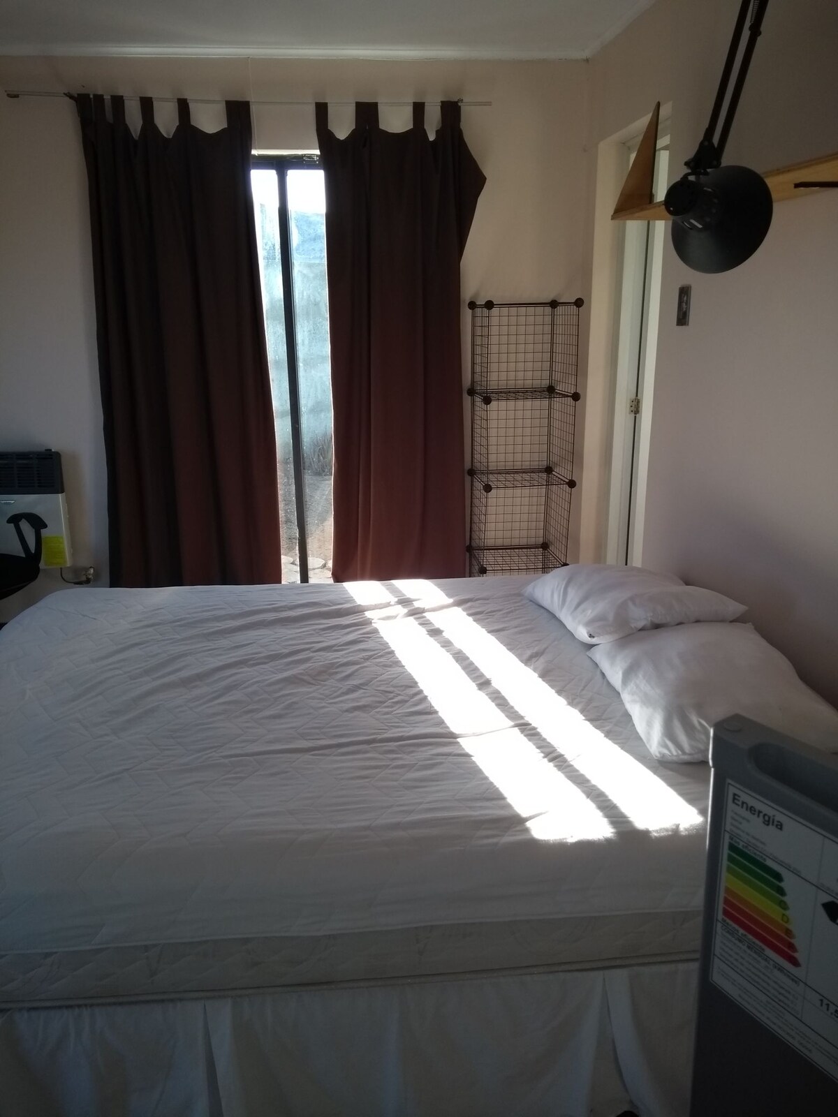 A bright sleeping area is seen, featuring a spacious bed with two pillows. Natural light filters through a sliding glass door, casting lines across the white bedding. Dark curtains frame the window, adding contrast to the light interior.