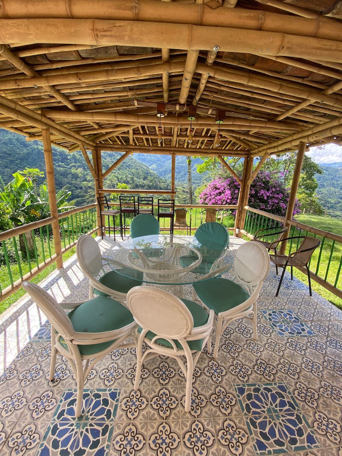A spacious outdoor area features a circular glass table surrounded by six cushioned chairs, set against a backdrop of lush greenery and distant mountains. A bamboo structure supports a thatched roof, with vibrant purple flowers enhancing the natural scenery.