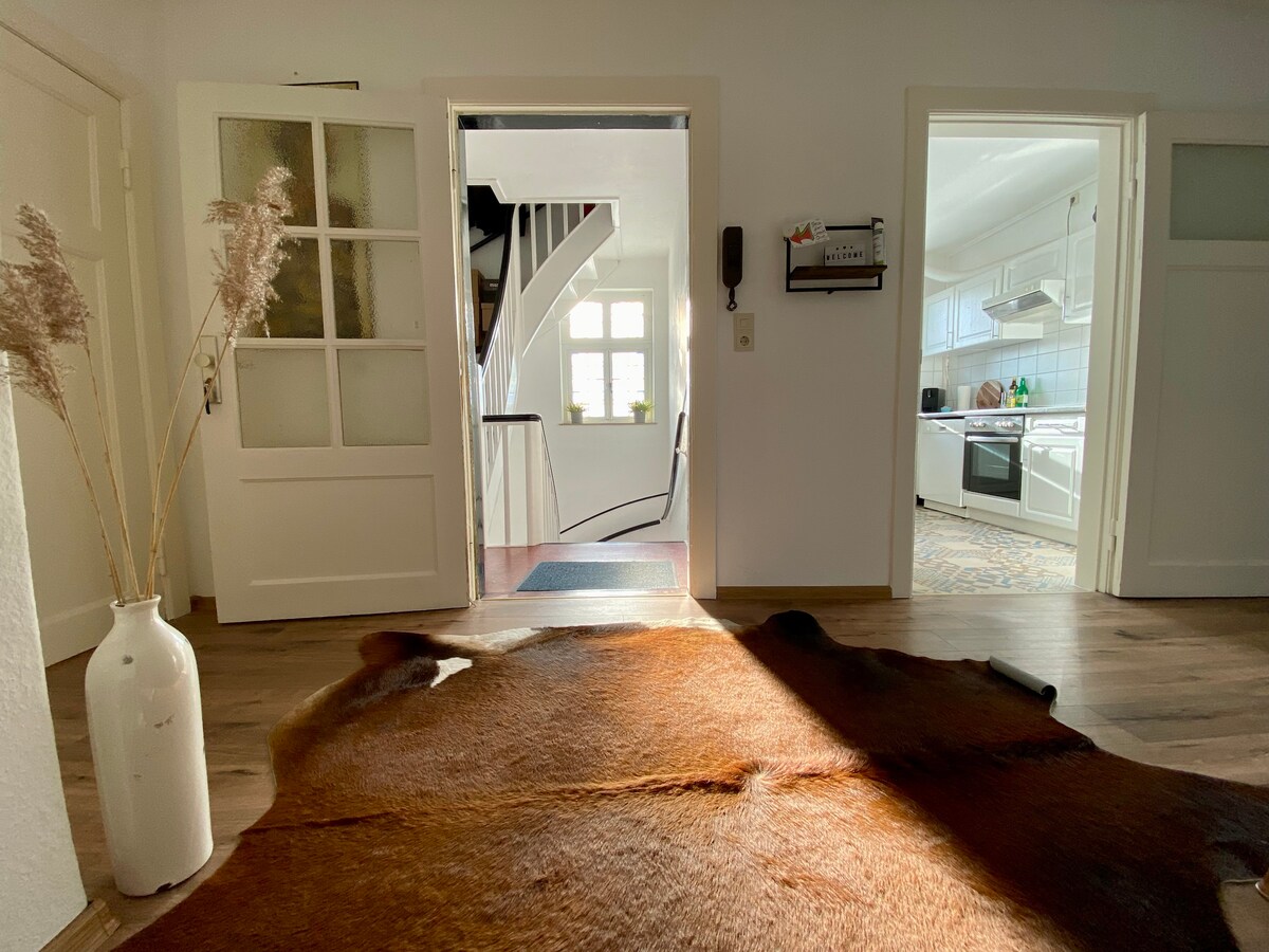 The entrance area features a warm-toned cowhide rug laid on the wooden floor. A white vase with dried pampas grass is positioned beside the door. Light enters through the glass-panel doors, illuminating the hallway leading to the kitchen and staircase.