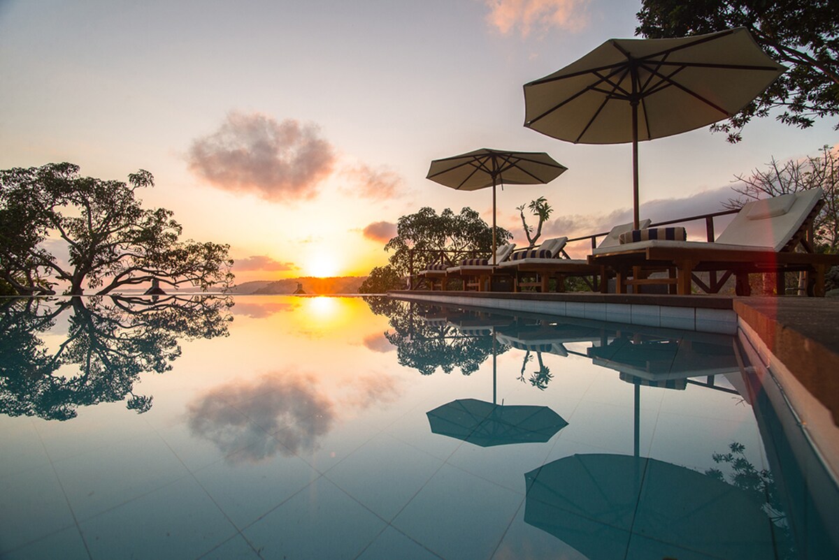 A serene pool area is depicted at sunrise, with the sky transitioning through soft hues. Sun loungers topped with umbrellas are positioned beside the water, while the reflective pool creates a tranquil visual experience of the surrounding nature.