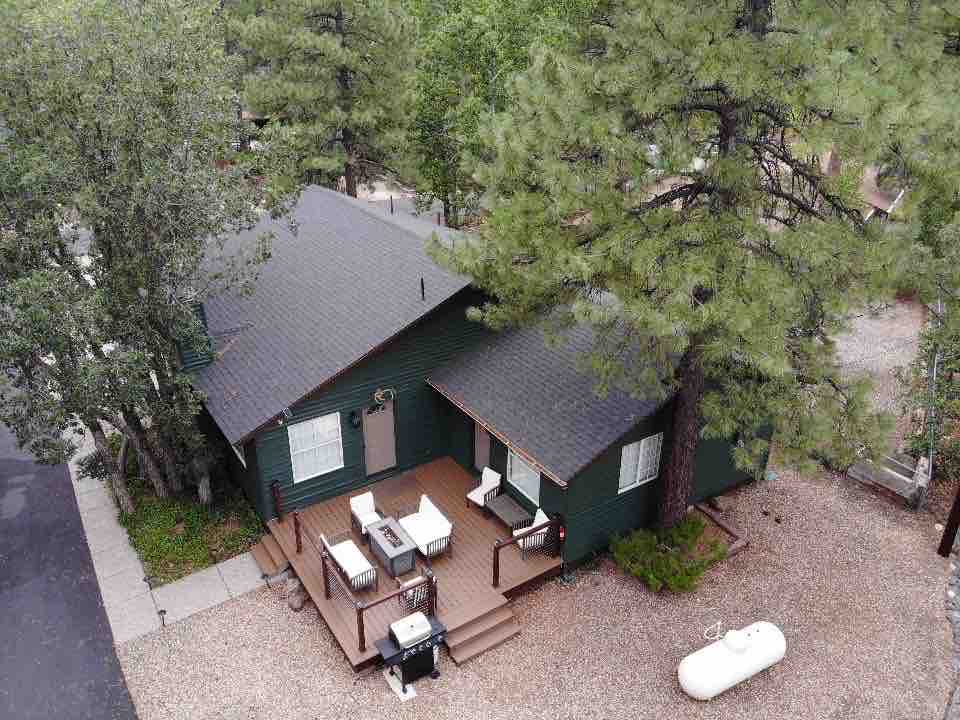 An aerial view of a cozy cabin reveals a green exterior surrounded by trees. A back deck features outdoor seating and a grill, situated on a gravel area. The cabin displays multiple windows, allowing natural light to enter the living space.
