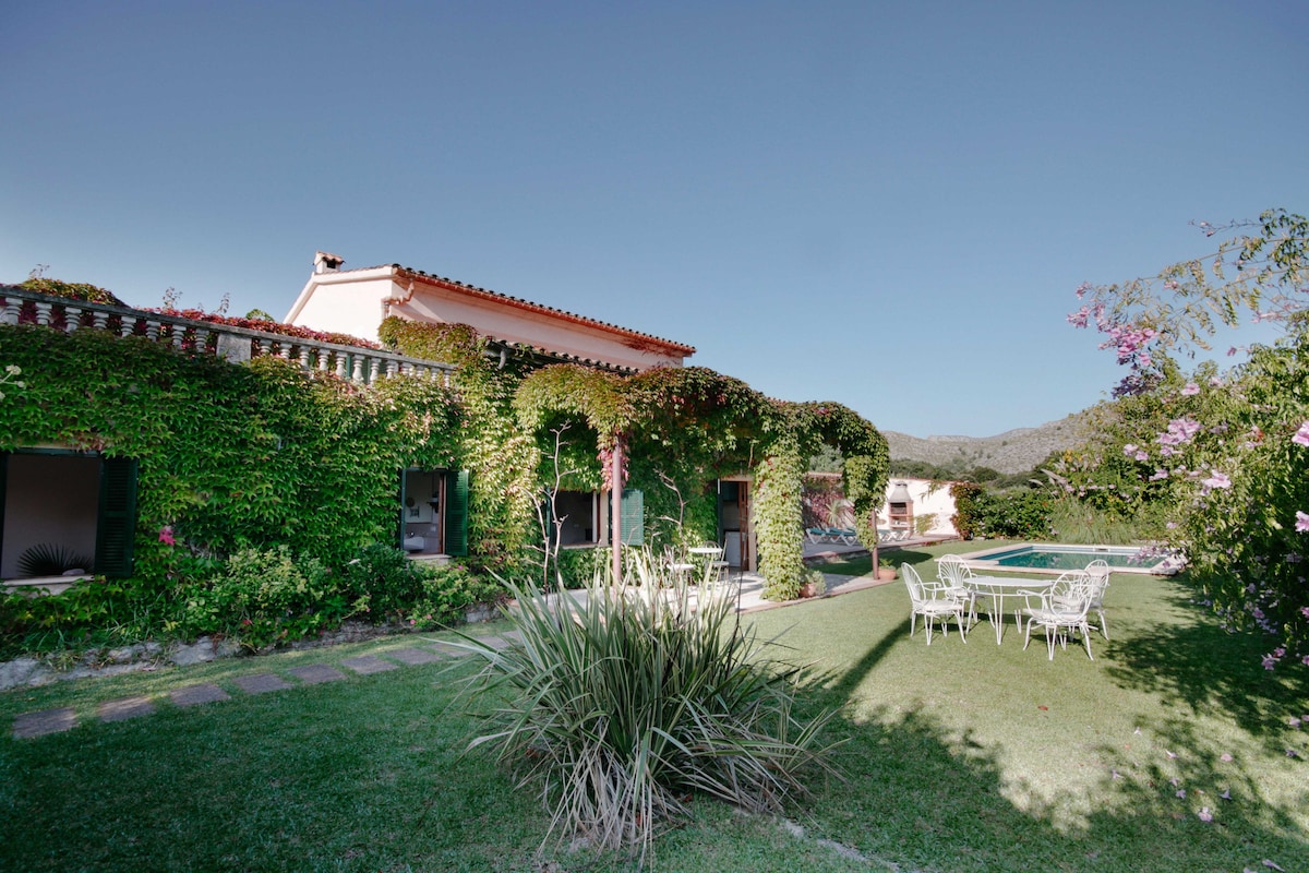 The exterior of the house is surrounded by lush greenery, featuring vibrant climbing plants covering the walls. A spacious garden area includes a swimming pool and a white outdoor dining set, creating an inviting space for relaxation amid the natural landscape.