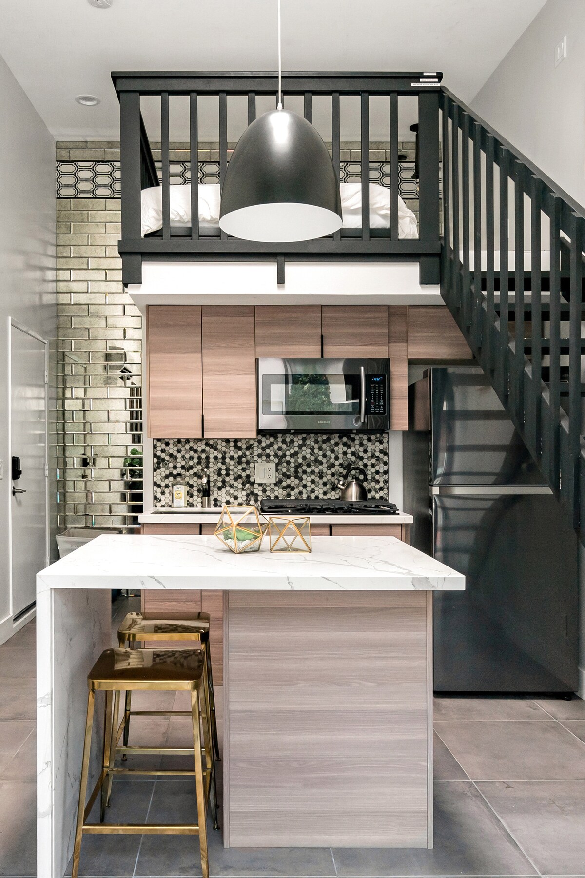 An open-concept kitchen is showcased, featuring modern cabinetry and a marble countertop. High stools are positioned along the island, and a stylish pendant light hangs above. The kitchen includes contemporary appliances and a mosaic tile backsplash, with a staircase leading to a lofted sleeping area.