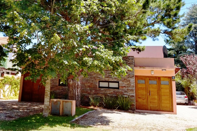 The exterior showcases a natural stone facade with wooden doors. A large tree provides shade in the front yard, while a gravel pathway leads to the entrance. A satellite dish is visible on the roof, and surrounding landscaped areas enhance the inviting setting.