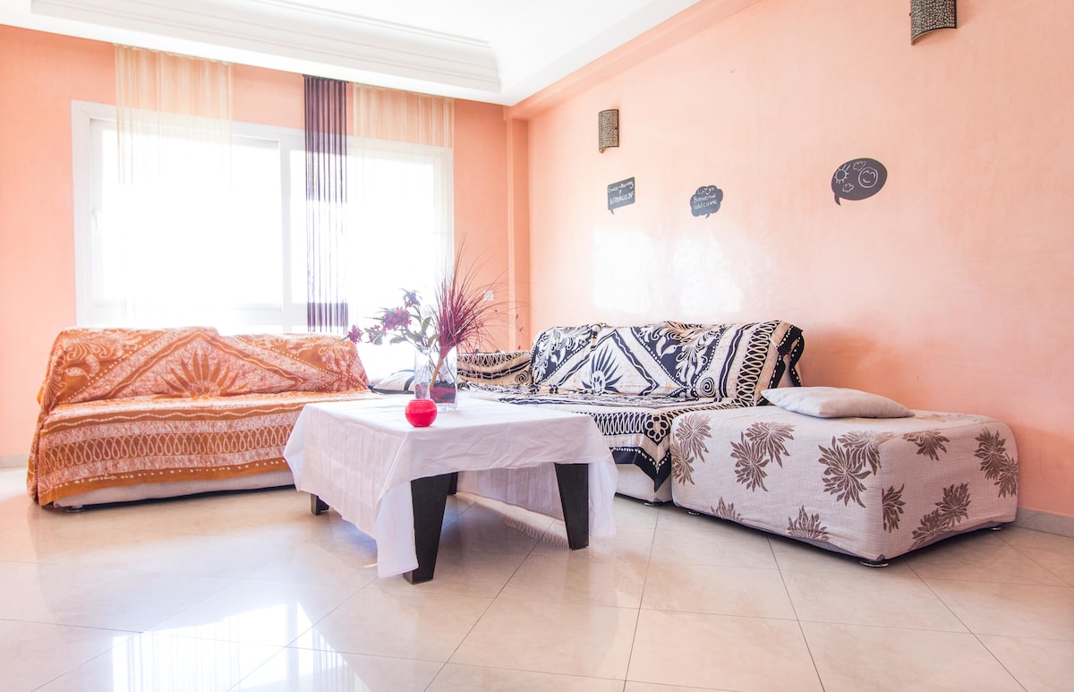 A bright living area features two patterned sofas draped with vibrant throws, facing a modest coffee table. Natural light fills the space through a large window adorned with sheer curtains, while decorative wall elements add character to the warm peach walls.