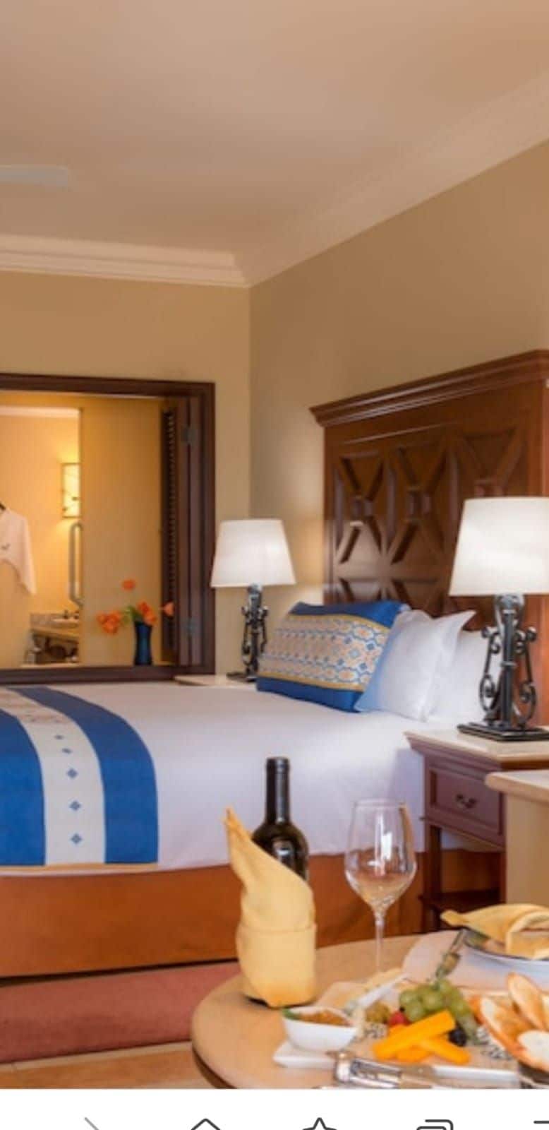 A well-furnished room features a spacious bed adorned with a blue and white bedspread. Two bedside lamps provide soft lighting on each side of the bed. The room includes a wooden dresser and a dining area displaying a bottle of wine and an assortment of food.