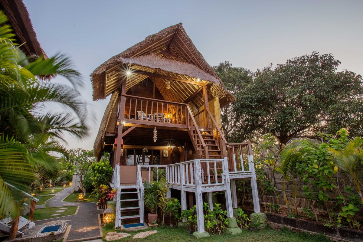 A charming two-story hut featuring a thatched roof is surrounded by lush greenery. Stairs lead up to a spacious balcony adorned with seating options, offering a welcoming outdoor space. Soft lighting highlights the garden paths, enhancing the tranquil atmosphere of the setting.