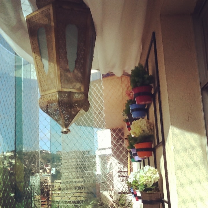 A decorative lantern hangs from the ceiling, casting soft shadows. Colorful flower pots line the wall, each featuring vibrant blooms in shades of white, blue, and pink. The transparent glass railing allows for an unobstructed view of the exterior, creating a light-filled atmosphere.