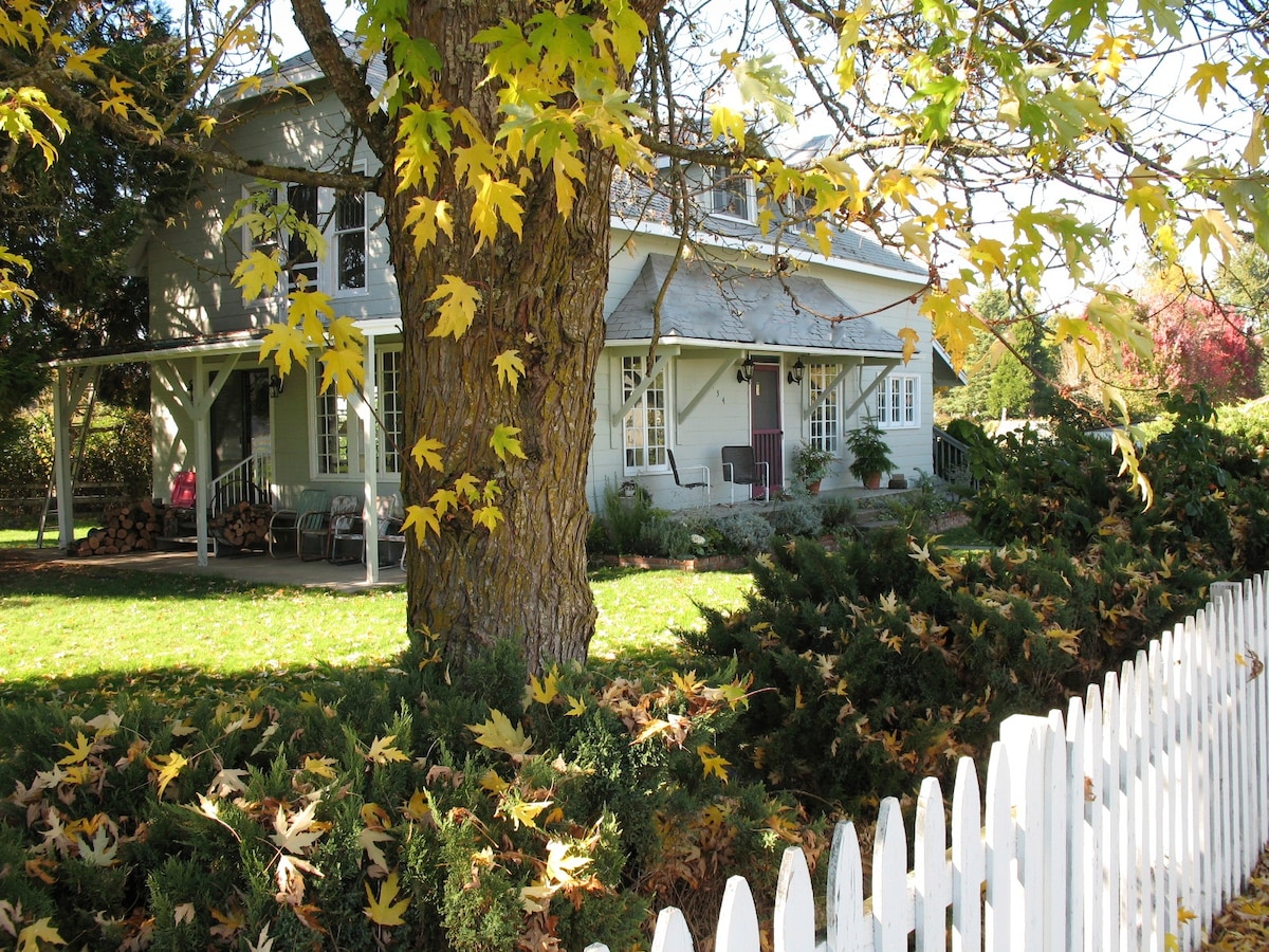 The exterior of a charming two-story farmhouse is framed by leafy trees in vibrant fall colors. A well-maintained white picket fence outlines the property, with a porch visible in front, featuring seating that invites relaxation amidst the peaceful garden setting.