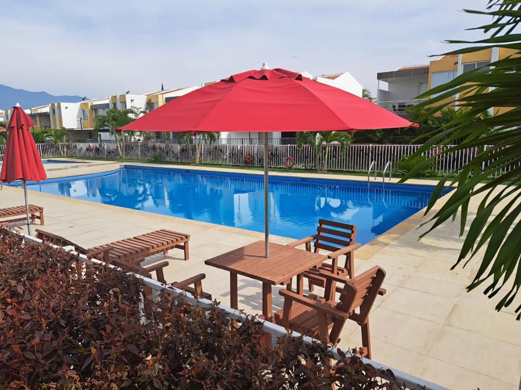 A large swimming pool is surrounded by a stone deck and adorned with vibrant red umbrellas. Several wooden lounge chairs are positioned nearby, offering shaded seating options. Lush greenery and a low hedge provide added privacy to the serene outdoor space.