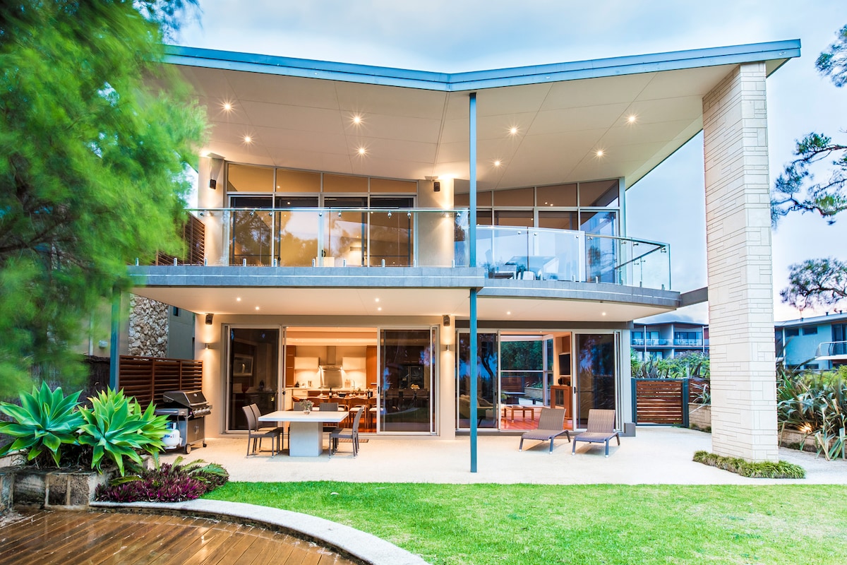 The exterior of a modern two-story house is displayed, featuring large glass doors that open to a spacious patio area. Comfortable outdoor seating is arranged around a dining table. Lush greenery surrounds the property, enhancing the connection between indoor and outdoor spaces.
