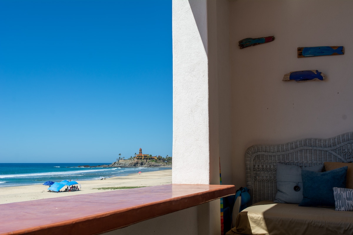 A view from the beachfront terrace reveals sandy shores and clear blue waters. Colorful umbrellas dot the beach, while a gentle wave pattern creates a serene backdrop. The terrace features a cozy day bed, inviting relaxation and enjoyment of the coastal scenery.