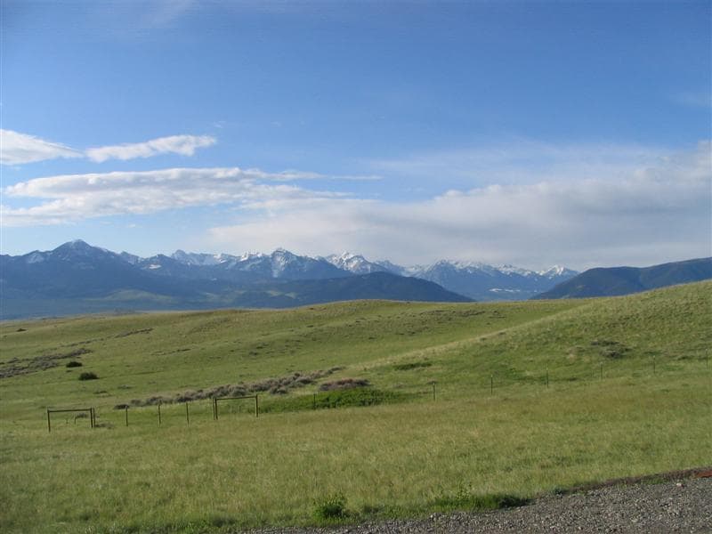 A panoramic view of the Absaroka Mountain range is displayed, showcasing snow-capped peaks under a clear blue sky. Lush green hills and patches of golden grass stretch toward the horizon, creating a sense of open space and tranquility.