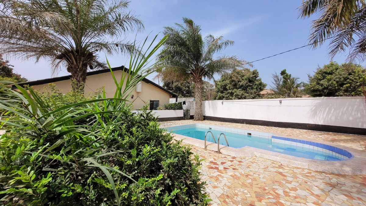 A serene communal pool is surrounded by lush greenery and palm trees, providing a relaxed atmosphere. The pool area features tiled edging and steps leading into the water, with a spacious layout allowing for leisurely enjoyment under the clear blue sky.