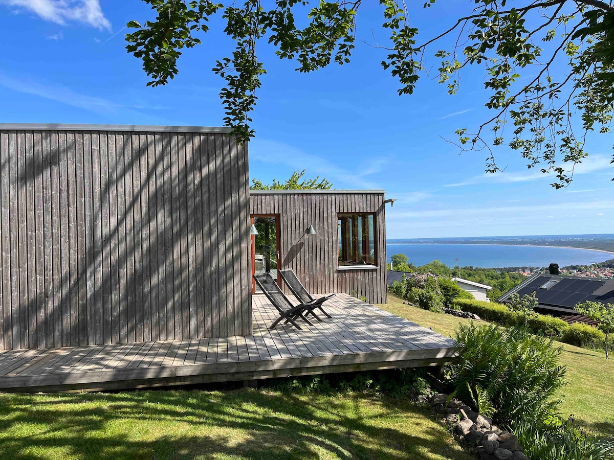 A contemporary wooden house stands on a wooden deck, surrounded by lush greenery. Two lounge chairs are positioned for scenic views of the coastline and the town below, with clear skies providing ample natural light.