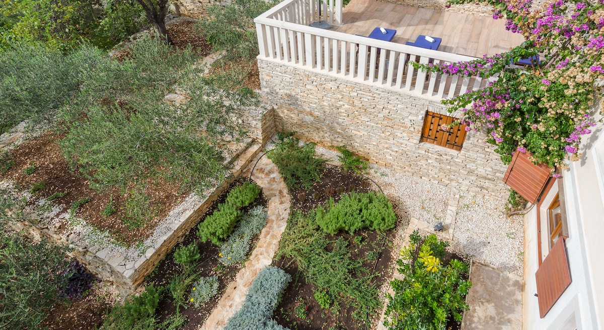 A bird's-eye view reveals a vibrant garden featuring rows of greenery and herbs, bordered by a stone pathway. An outdoor terrace is visible above, complete with brown loungers, while climbing plants adorn the stone wall, providing a touch of natural color.