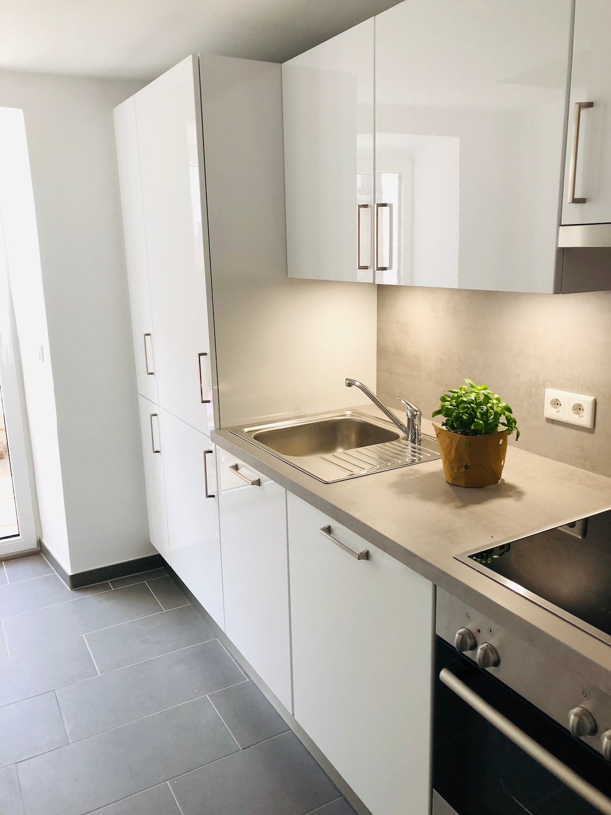A modern kitchen is presented with sleek white cabinetry and a stainless steel countertop. A small potted plant adds a touch of greenery. Natural light enters through a nearby window, enhancing the clean and organized space.