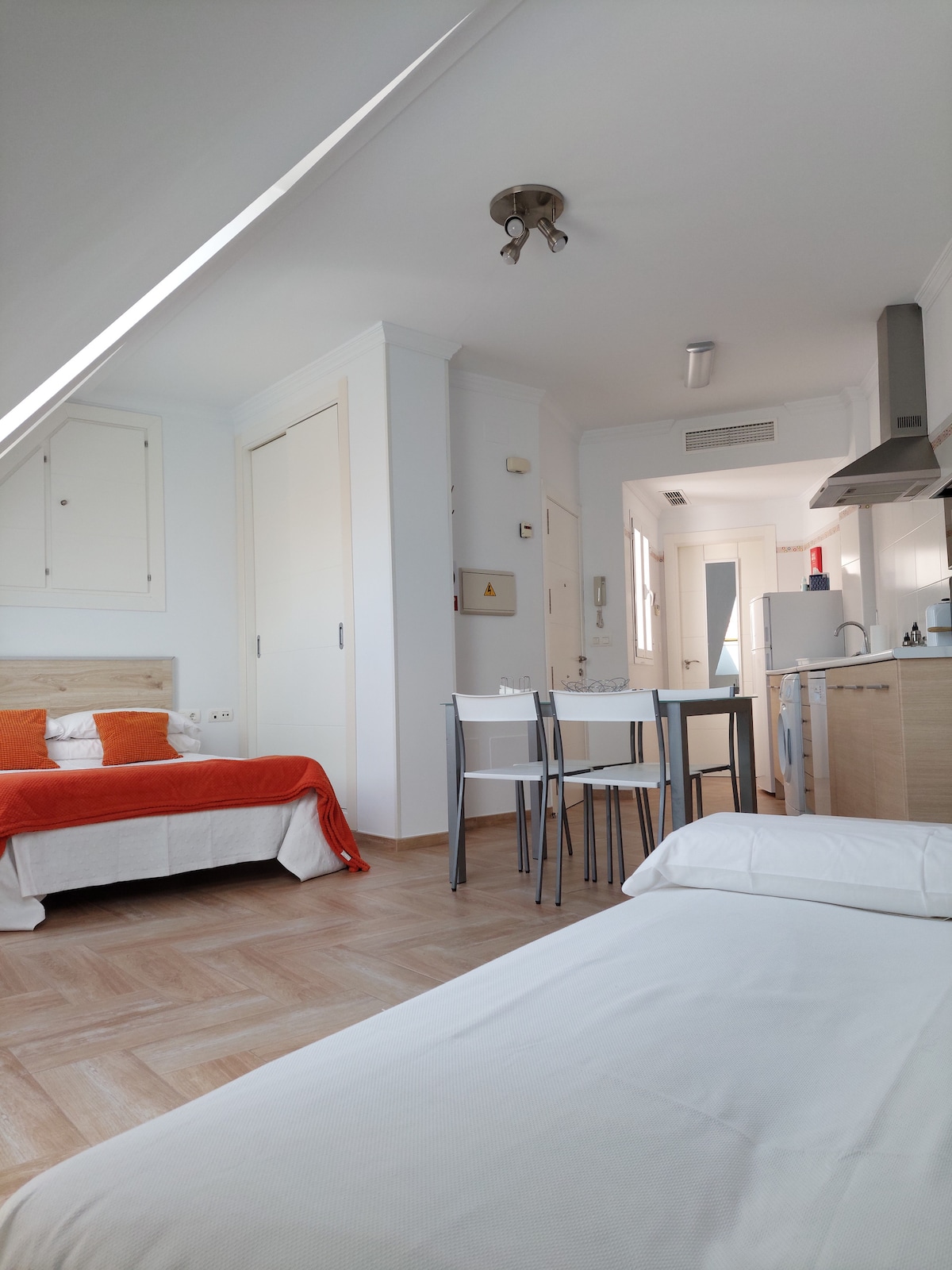 A bright studio space features a comfortable double bed dressed in white linens and an orange throw blanket. A dining table with four chairs is positioned near the kitchen, equipped with modern appliances. Light wood cabinetry complements the overall airy atmosphere.