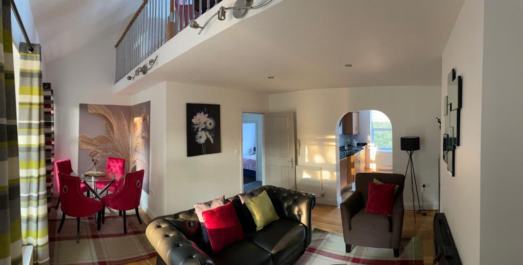 An open-plan living area features a comfortable dark leather sofa adorned with vibrant cushions. A dining table with two red chairs is positioned nearby, while soft lighting enhances the space. An archway leads to the kitchen area, visible in the background.