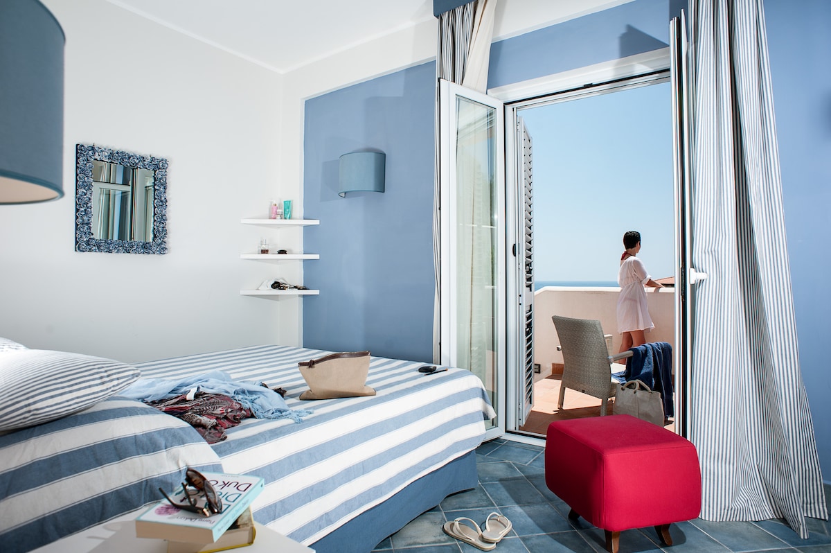 A bright bedroom is showcased with blue and white striped bedding on a spacious bed. A cozy red ottoman complements the space. Sunlight filters through the glass doors that lead to a private balcony, where a person can be seen enjoying the view.