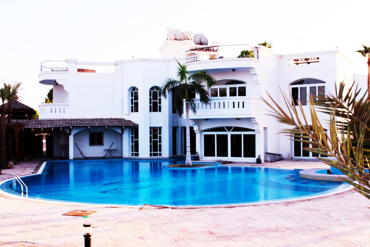 The exterior of a white villa features large windows reflecting natural light. A spacious pool is seen in the foreground, surrounded by palms and a tiled deck. The structure showcases a combination of architectural styles with multiple balconies and an inviting entrance.