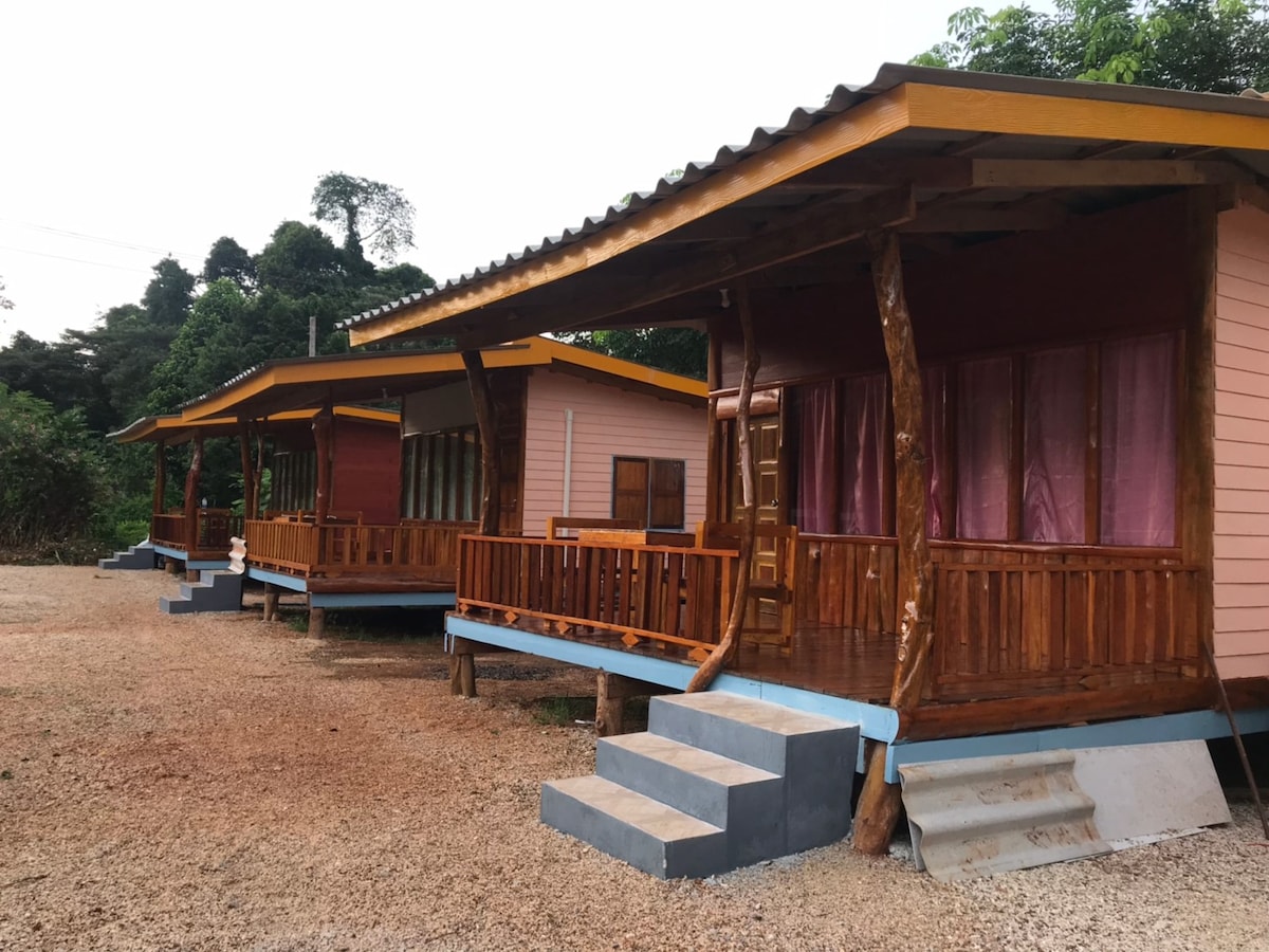 A row of colorful bungalows is set on a gravel pathway, featuring wooden verandas with railings. Each bungalow showcases large windows with pink curtains, allowing natural light to fill the interior. The structure is surrounded by lush greenery, creating a serene atmosphere.
