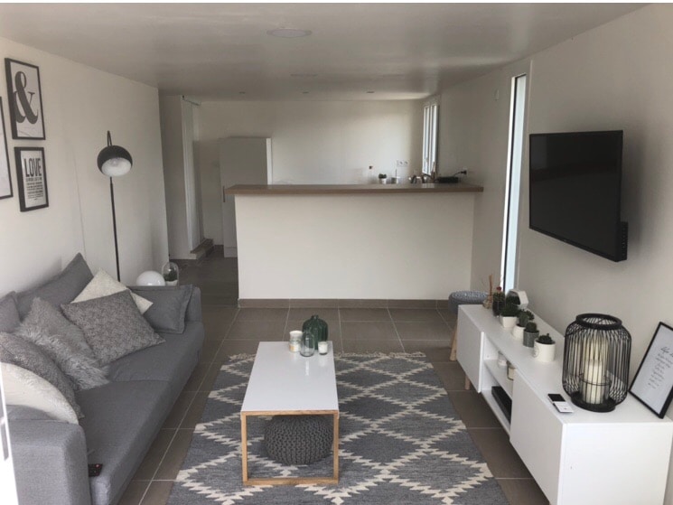 A spacious living area is displayed, featuring a plush gray sofa with decorative cushions. A television is mounted on the wall opposite a minimalist white entertainment unit. A coffee table sits on a patterned rug, while a modern lamp and decorative items add a touch of style.
