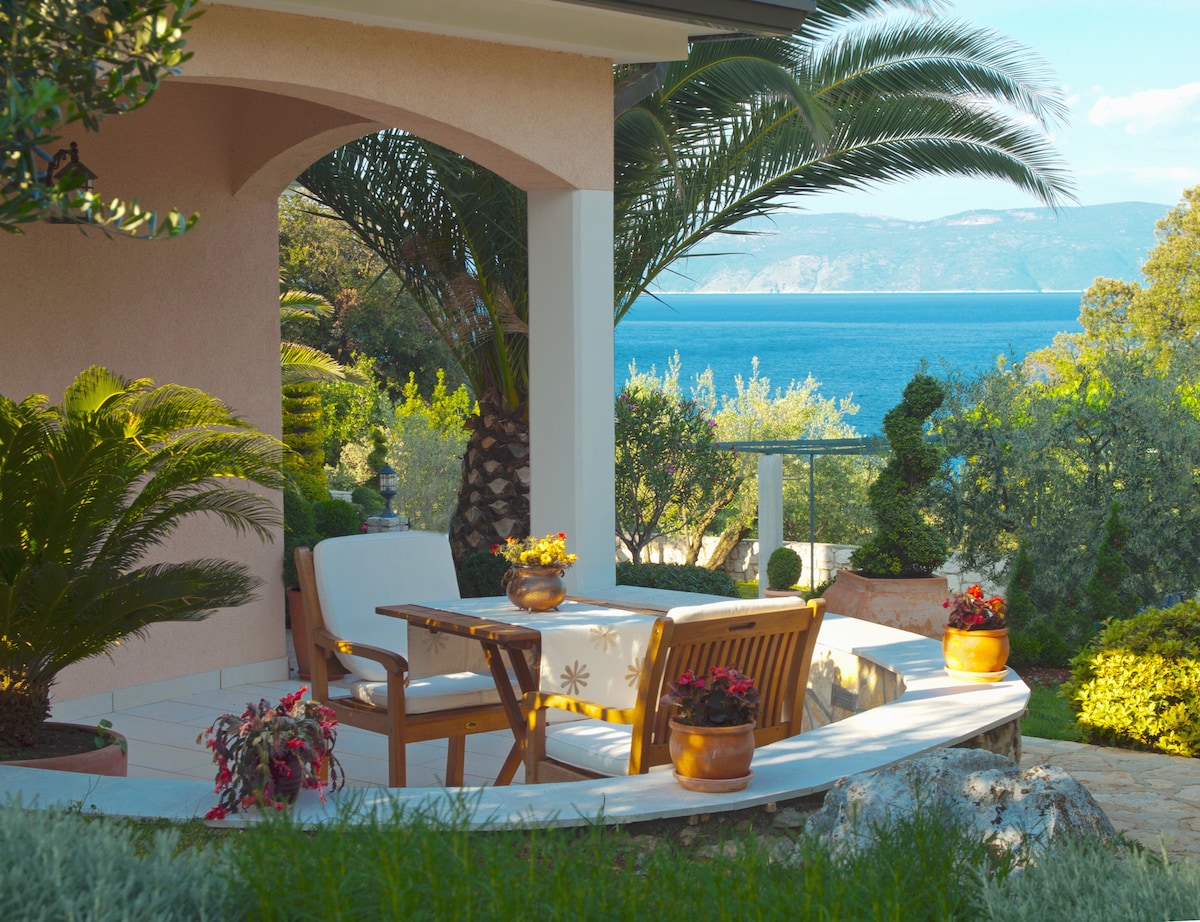 An outdoor seating area features a wooden table with two chairs, surrounded by potted flowers and lush greenery. Palm trees sway in the gentle breeze, creating a tranquil atmosphere, while the expansive view of the sea and distant hills enhances the serene setting.