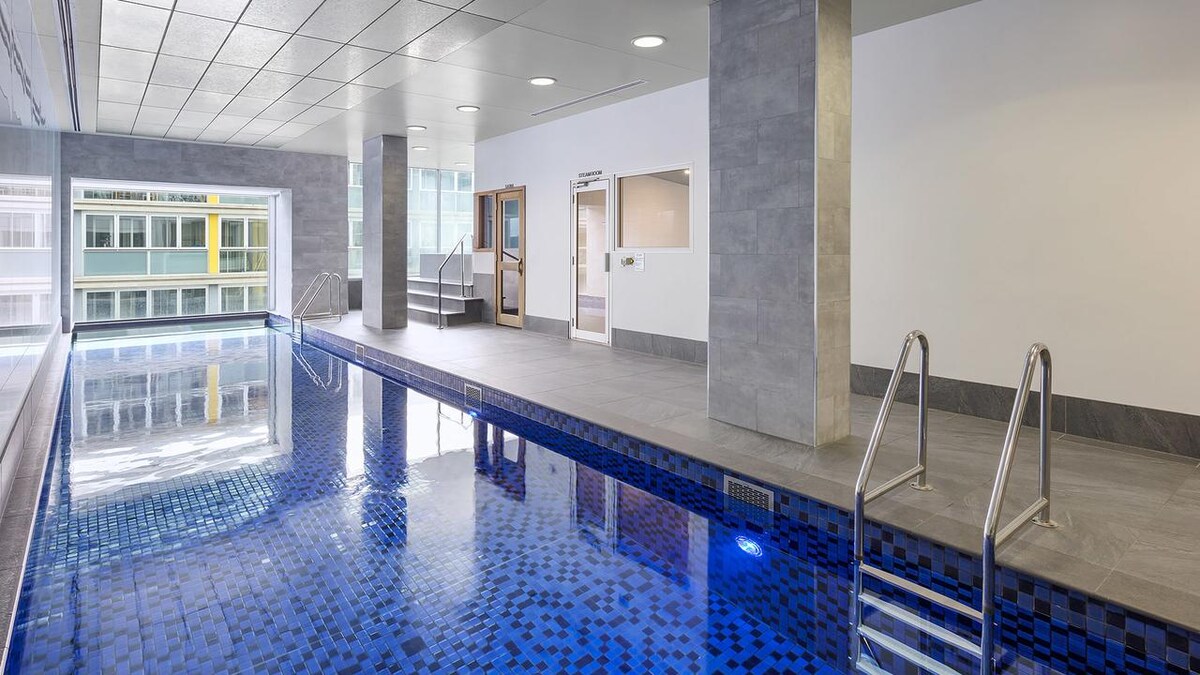 A sleek indoor pool area features a clear blue water surface and modern tile accents. Lighting highlights the spacious layout, complemented by large windows that allow natural light to fill the space. Two stairways provide access to the pool, enhancing the inviting atmosphere.