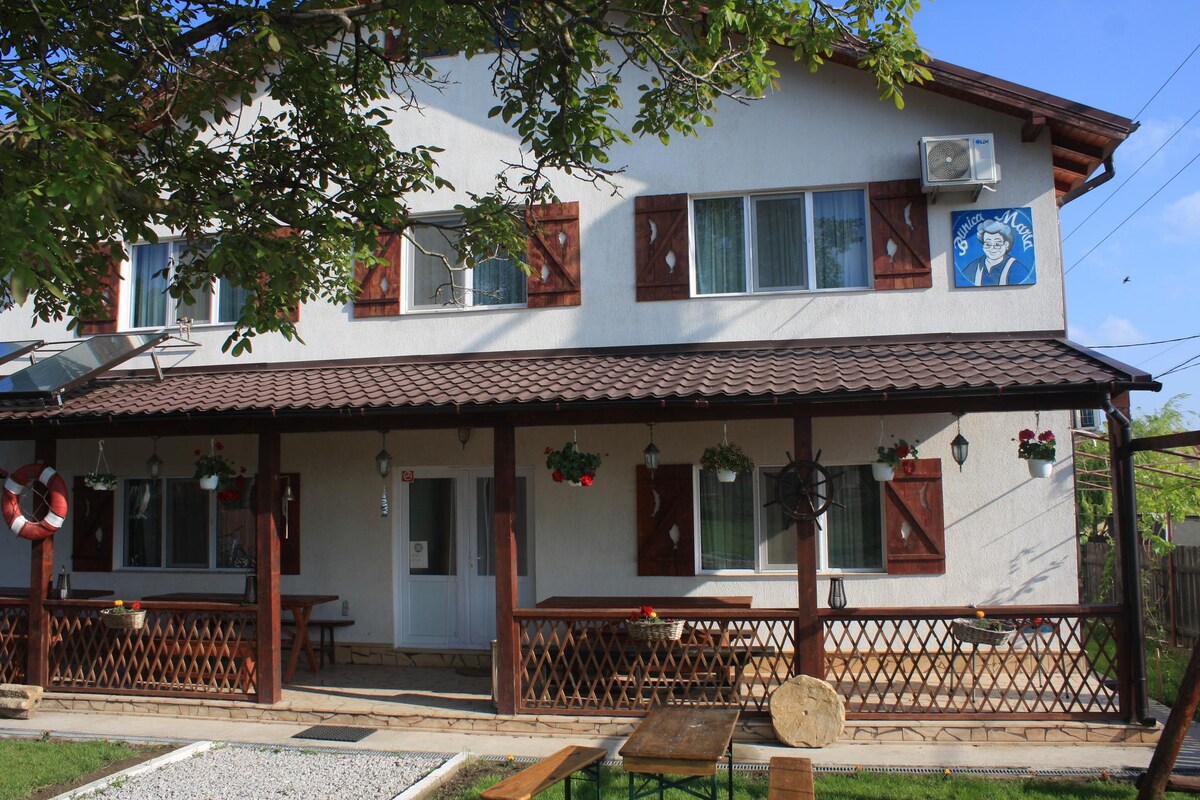 The guesthouse exterior features a two-story white structure with brown shutters and a tiled roof. A shaded porch is adorned with hanging plants and wooden tables. A gravel path leads to the entrance, surrounded by greenery, enhancing the welcoming atmosphere of the accommodation.