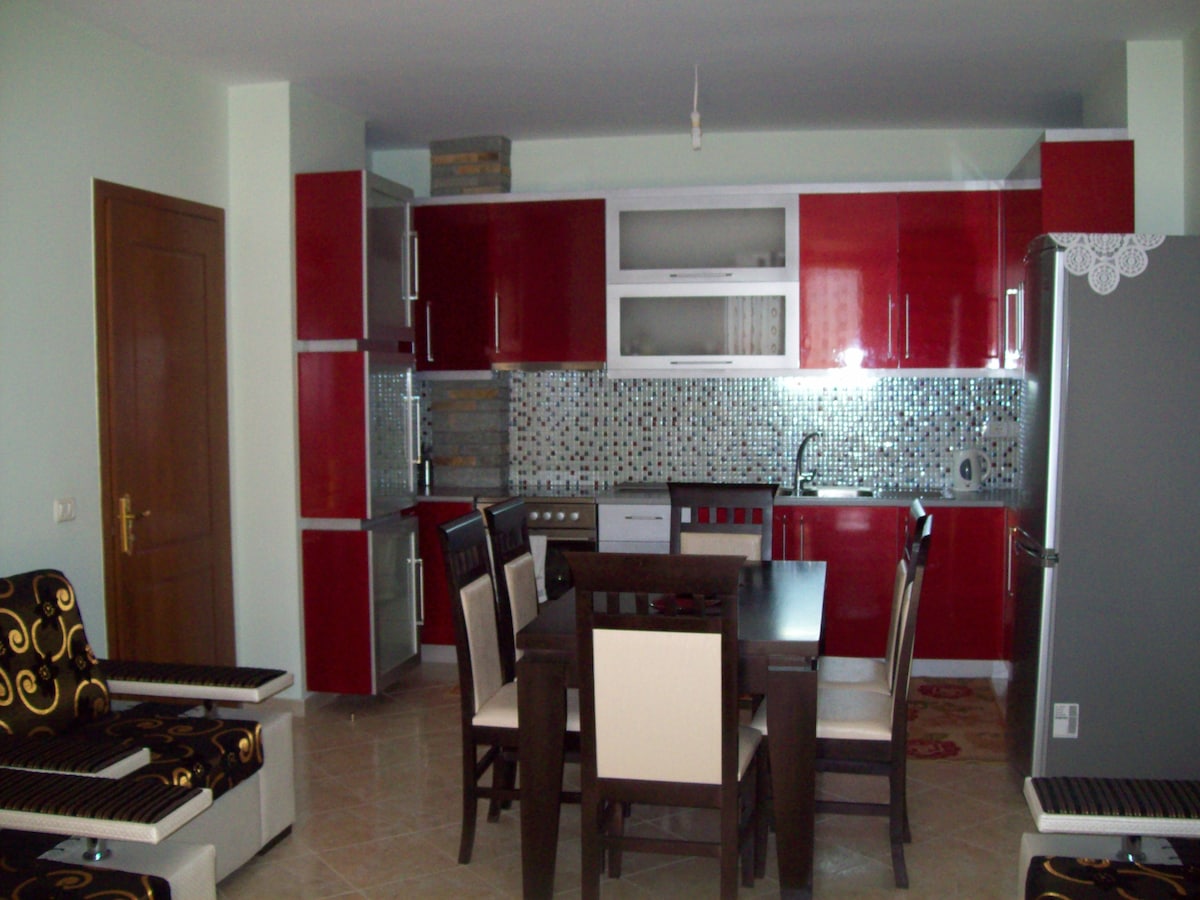 A modern kitchen and dining area are presented, featuring sleek red cabinetry and a mosaic-tiled backsplash. A dining table with contrasting chairs is accompanied by a refrigerator and a well-equipped cooking area. Natural light brightens the space, creating an inviting atmosphere.