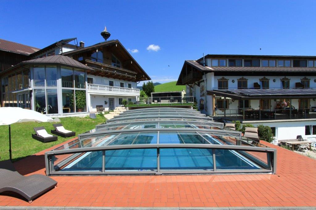 An outdoor heated pool is featured with a glass cover, providing a clear view of the surrounding landscape. Sun loungers line the deck, and the backdrop includes spacious buildings with traditional architecture, set against a vibrant blue sky.