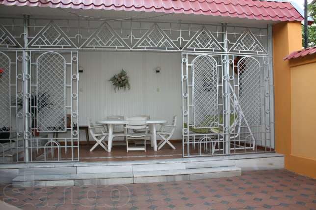 A covered outdoor patio area features a set of white wooden furniture, including a table and chairs. Decorative grill work frames the space, providing an open yet secure environment. The floor is tiled with a geometric pattern, enhancing the inviting atmosphere.