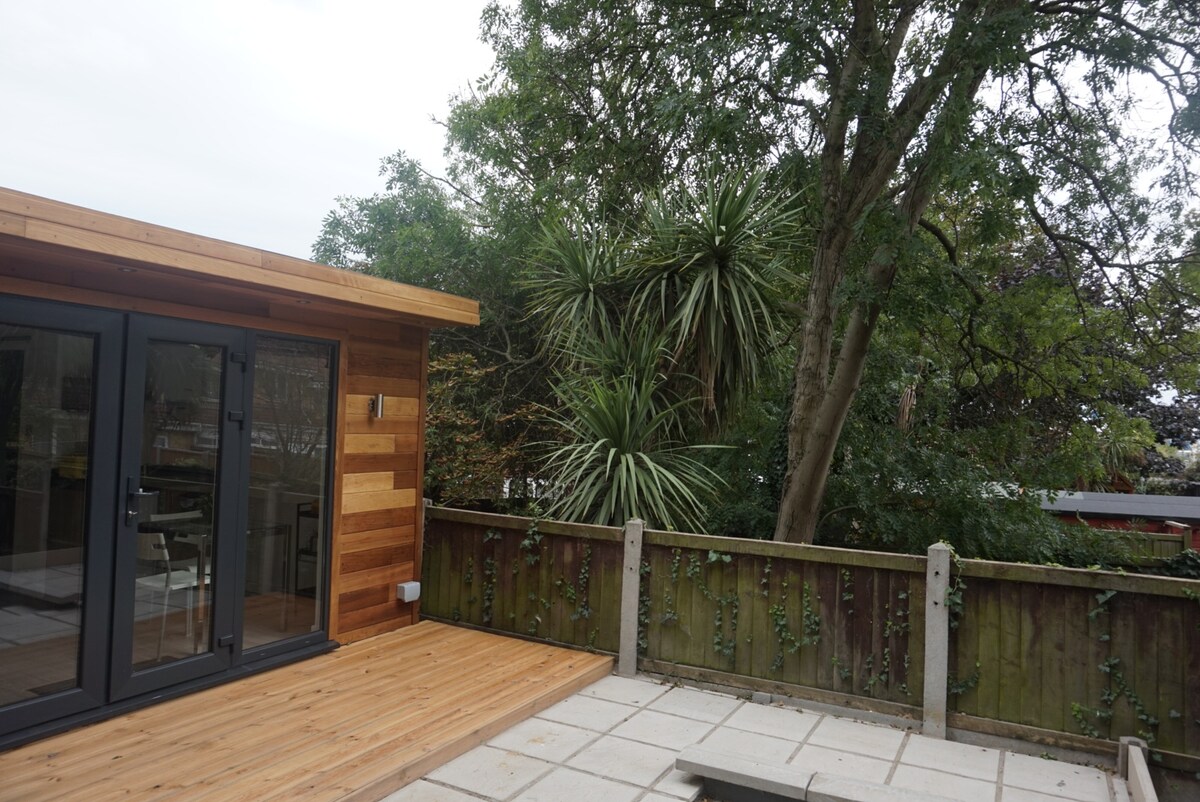 A wooden deck area is shown, bordered by a fence covered in greenery. The modern structure features large glass doors that open to the outside. Lush trees and palm-like plants add a natural touch to the relaxing outdoor space.