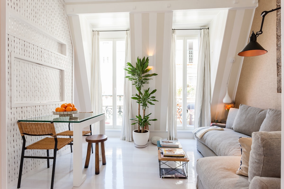 A bright living area features a comfortable grey sofa and a glass dining table accompanied by two woven chairs. Natural light filters through tall windows adorned with sheer curtains, illuminating a potted plant that adds a touch of greenery to the space.
