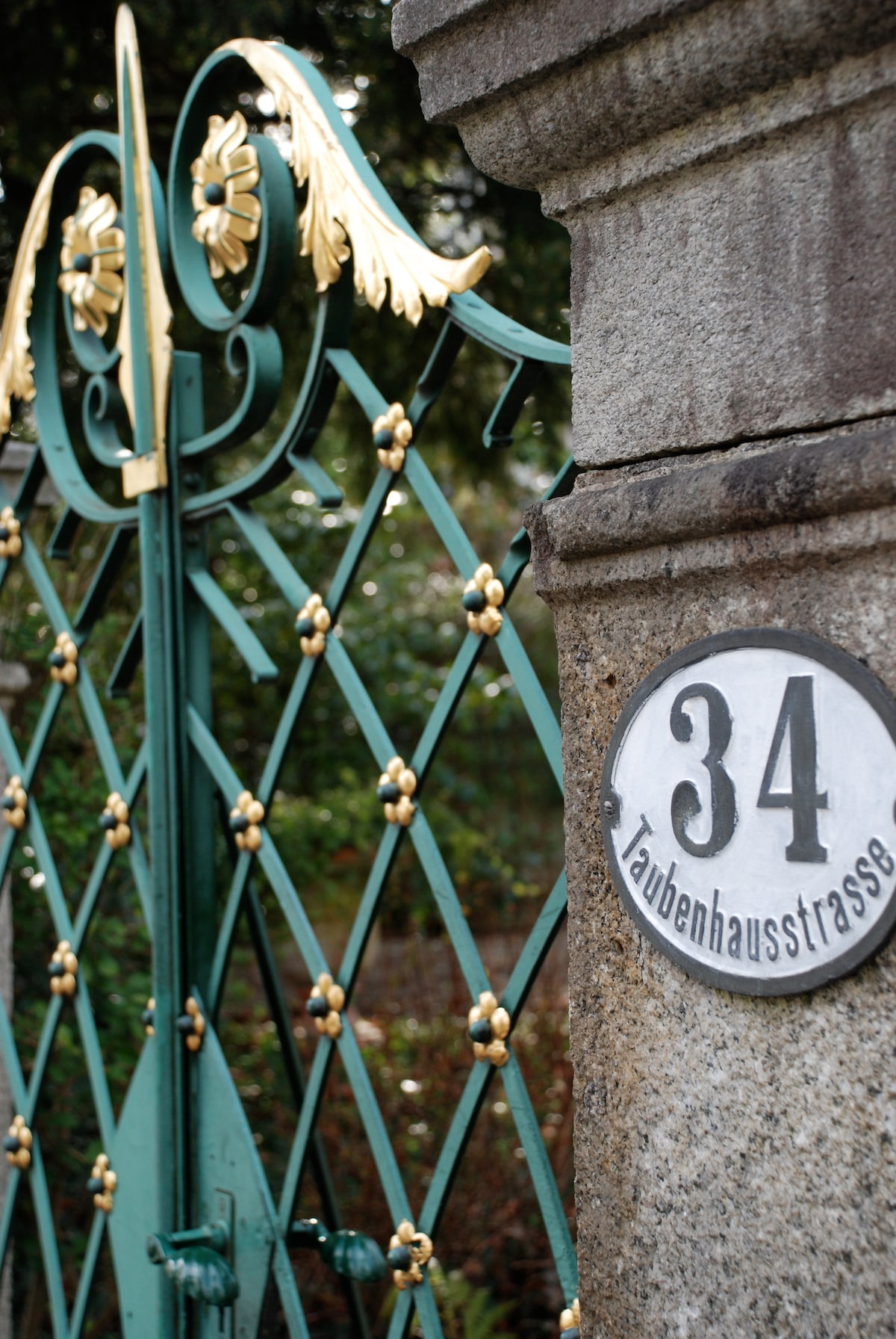 An ornate green gate features intricate metalwork with golden accents. The gate is positioned next to a stone pillar displaying the house number 34, indicating the entrance to the property. Surrounding foliage is softly blurred in the background.
