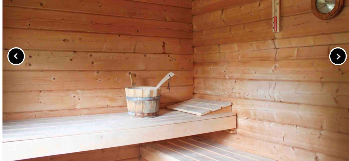 The sauna interior features light wood paneling, creating a warm environment. Wooden benches are positioned along the walls, with a wooden bucket nearby. A small shelf rests against one wall, and a thermometer is mounted for temperature monitoring.