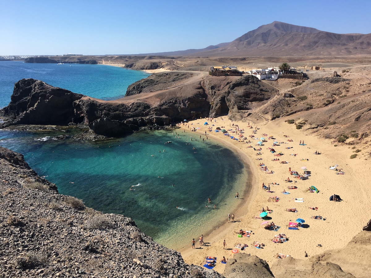 A vibrant beach scene features soft golden sand surrounded by rugged cliffs. Turquoise waters gently lap at the shore, while sunbathers and colorful beach umbrellas dot the landscape. The background showcases a coastal town nestled against a mountainous backdrop.