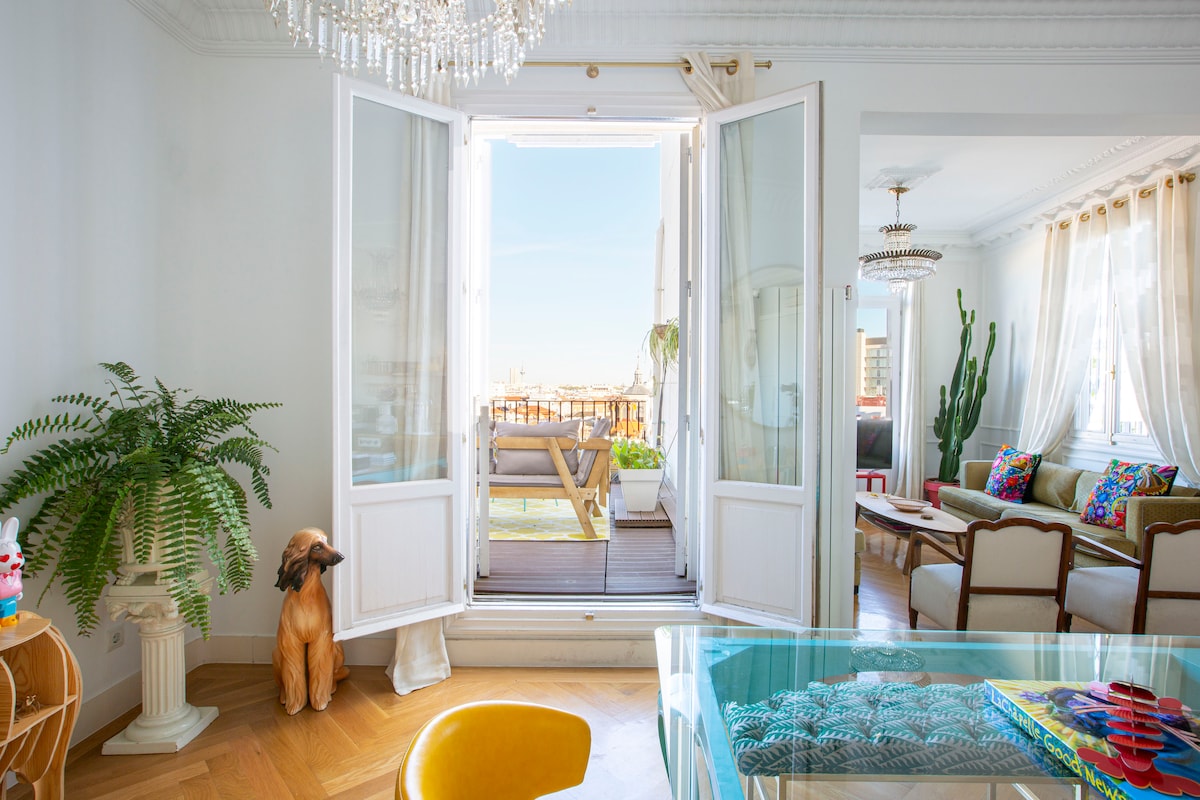 Bright living area featuring an open balcony with views of the city. Large glass doors lead to a sunny terrace. A decorative dog sculpture and potted plants add a touch of nature, while a dining table with a colorful centerpiece is visible in the foreground.