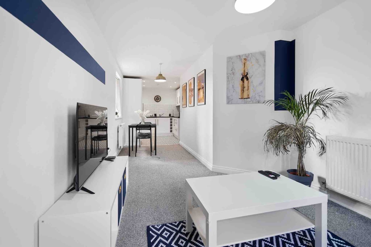 A bright living area is featured with a modern white coffee table and a flat-screen TV. A potted plant adds a touch of greenery. Walls are adorned with framed art pieces, and a dining space is visible in the background, featuring a small table and chairs.