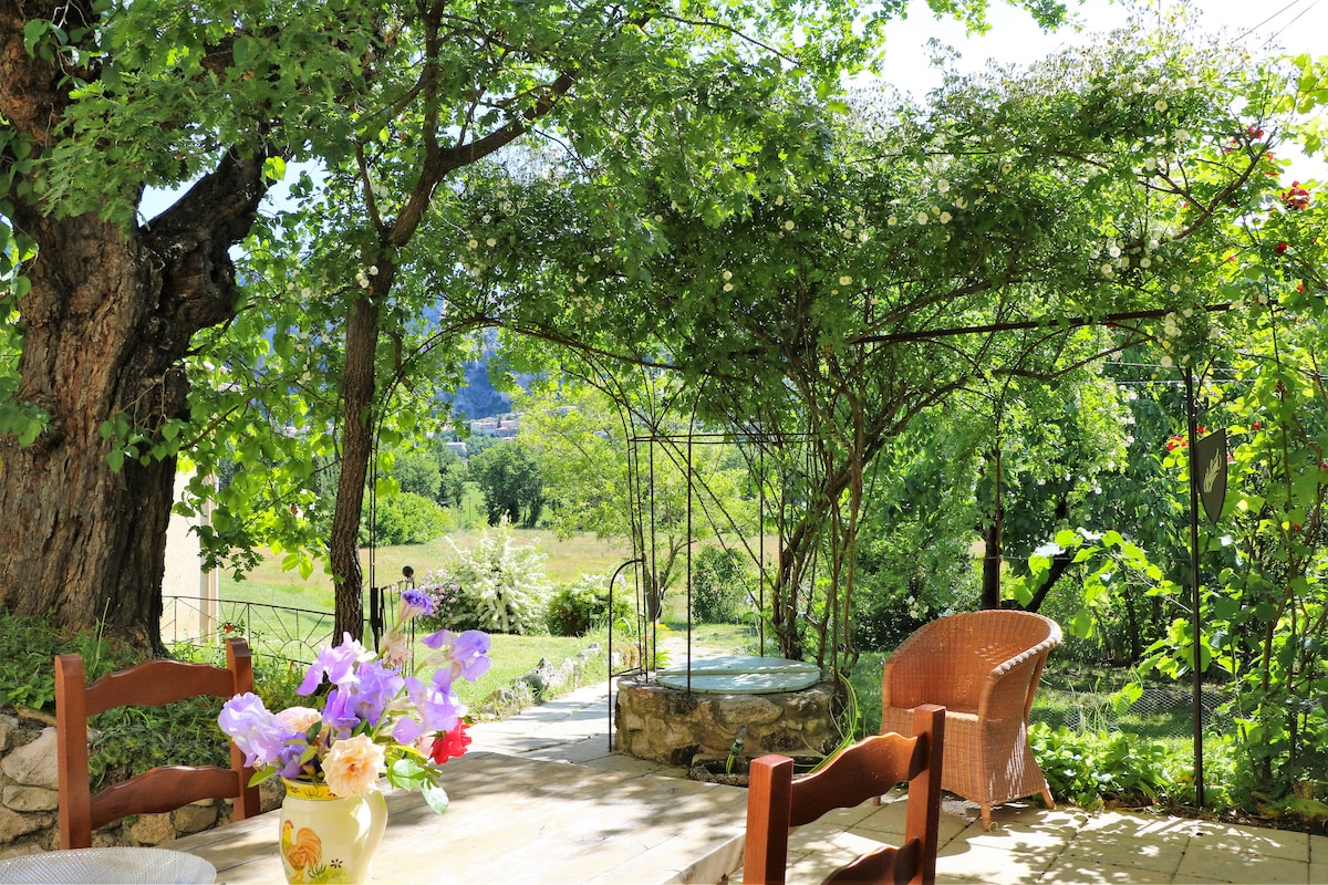 A shaded outdoor seating area is framed by lush greenery and trees. A wooden table, adorned with a vibrant bouquet of flowers, is positioned alongside a stone feature. A comfortable wicker chair is visible, inviting relaxation in a serene garden setting.