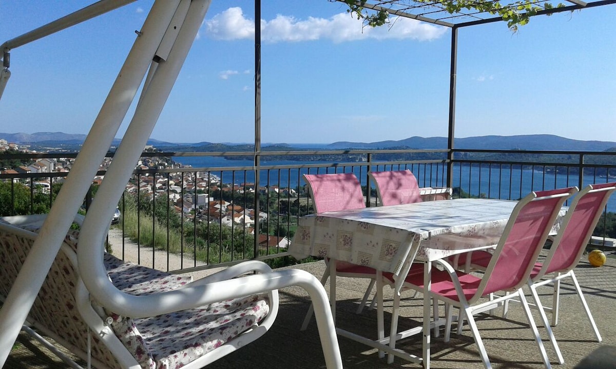 A large terrace features a table surrounded by several pink chairs, offering a scenic view of the city and nearby islands. Two swings are positioned nearby, with a railing providing safety and an open sky overhead. The serene landscape is framed by distant hills and water.