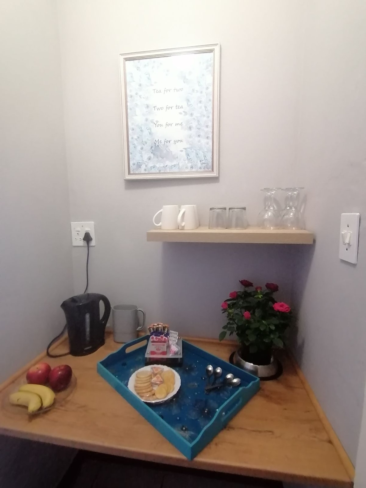 A small kitchenette corner is presented with a wooden shelf displaying two glasses and a teapot. A blue tray holds an assortment of snacks, with a plate of cookies, silverware, and fruits below. A small potted plant adds a touch of greenery.